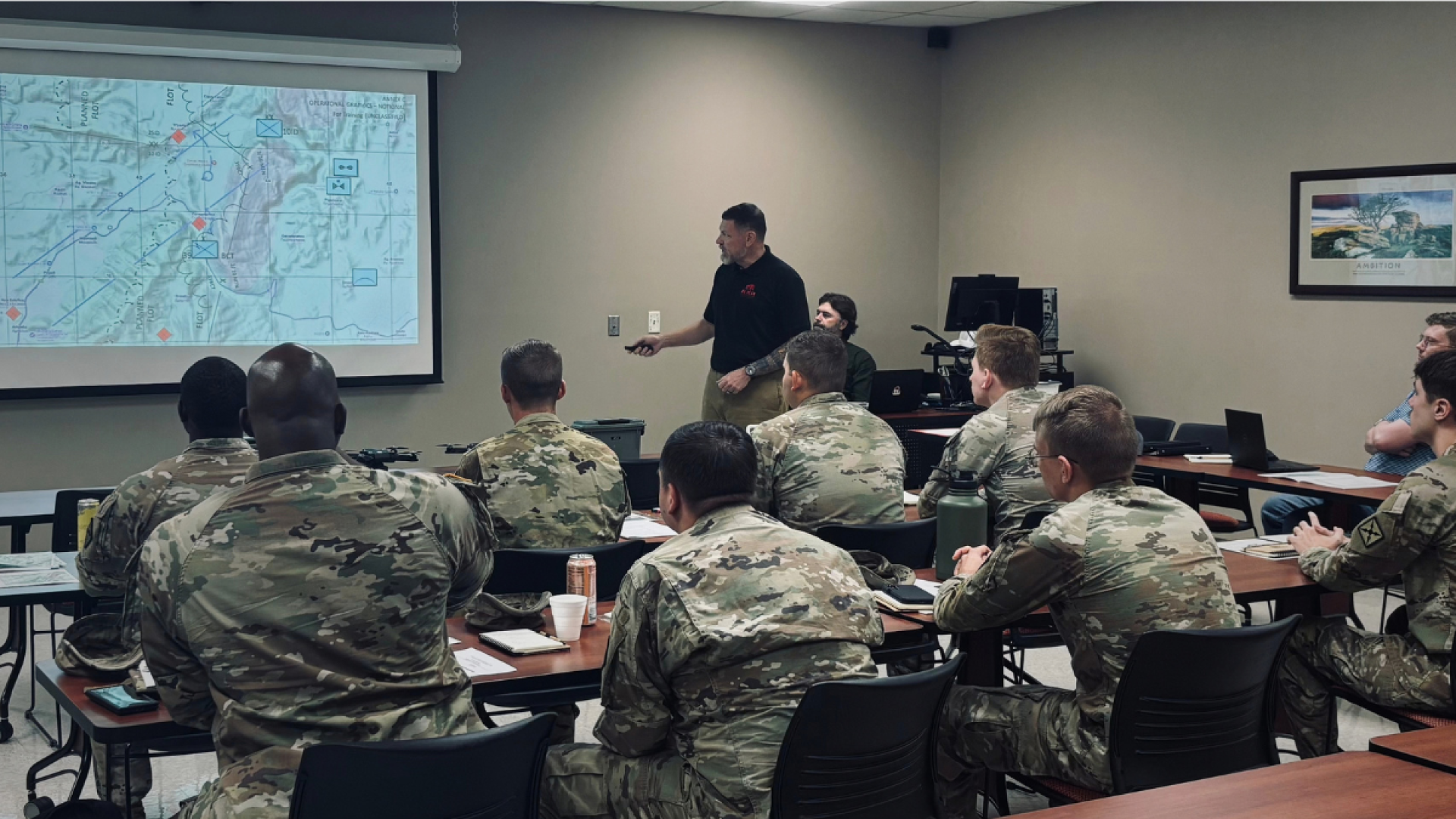 An instructor presents a tactical map on a projector screen to a classroom of Arkansas Army National Guardsmen in camouflage uniforms seated at tables.